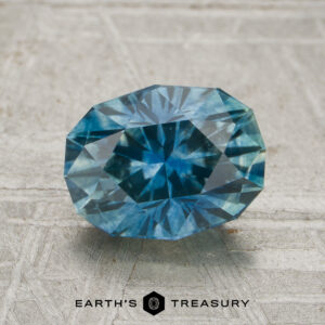 A 1.76-carat teal blue Montana sapphire (heated), cut in an oval shape, is displayed on a textured gray surface above the text “EARTH’S TREASURY.”.