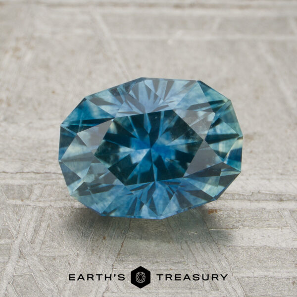 1.76-Carat Teal Blue Montana Sapphire (Heated) A 1.76-carat teal blue Montana sapphire (heated), cut in an oval shape, is displayed on a textured gray surface above the text “EARTH’S TREASURY.”.