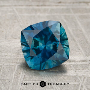A 1.80-carat cushion-cut Montana sapphire (heated) rests on a textured surface, with the “Earth’s Treasury” logo and text displayed below it.