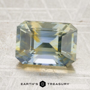 A rectangular, faceted 3.04-carat yellow-sky blue particolored Ceylon sapphire (heated) with vibrant blue, yellow, and green hues rests on a textured gray surface. The “EARTH’S TREASURY” logo appears at the bottom.
