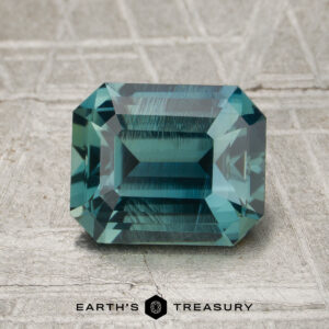 A rectangular emerald-cut 2.27-carat teal Madagascar sapphire rests on a textured gray surface, with the words “Earth’s Treasury” below it.