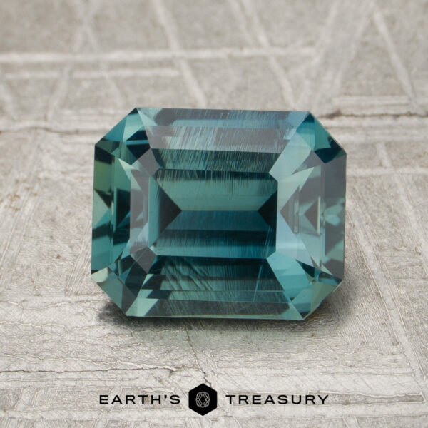 2.27-Carat Teal Madagascar Sapphire (Unheated) A rectangular emerald-cut 2.27-carat teal Madagascar sapphire rests on a textured gray surface, with the words “Earth’s Treasury” below it.