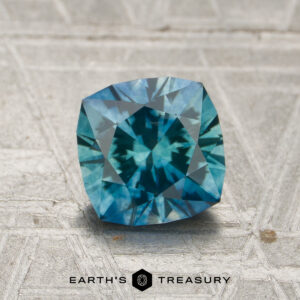 A 1.56-carat heated Montana sapphire is displayed on a textured surface, with the words “EARTH’S TREASURY” and a hexagonal logo beneath it.