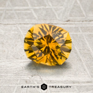 A 1.16-carat rich golden yellow Montana sapphire (heated) is displayed on a textured gray surface with the words “EARTH’S TREASURY” at the bottom.