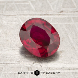 A 1.20-carat, oval-shaped faceted Mozambique ruby (heated) rests on a textured light surface, with the words “Earth’s Treasury” displayed at the bottom of the image.