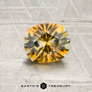 A 1.06-carat golden yellow–pale aqua particolored Montana sapphire with intricate faceting is displayed on a textured surface. The text “EARTH’S TREASURY” appears below the stone.
