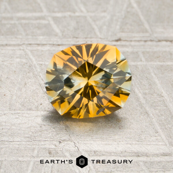 1.06-Carat Golden Yellow-Pale Aqua Particolored Montana Sapphire (Heated) A 1.06-carat golden yellow–pale aqua particolored Montana sapphire with intricate faceting is displayed on a textured surface. The text “EARTH’S TREASURY” appears below the stone.