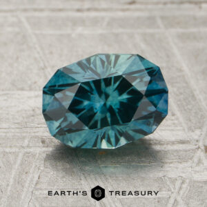 A 1.67-carat teal green-blue particolored Montana sapphire (heated) is displayed on a textured gray surface above the text “EARTH’S TREASURY” and a hexagonal logo.