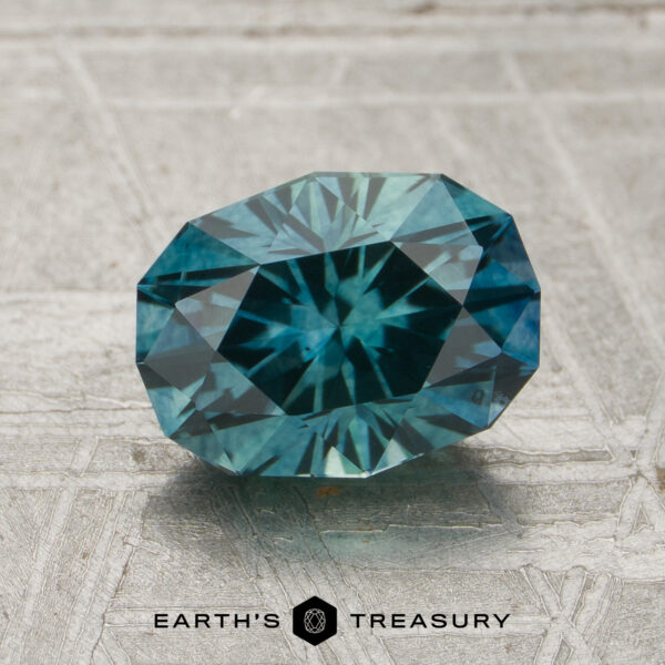 1.67-Carat Teal Montana Sapphire (Heated) A 1.67-carat teal green-blue particolored Montana sapphire (heated) is displayed on a textured gray surface above the text “EARTH’S TREASURY” and a hexagonal logo.