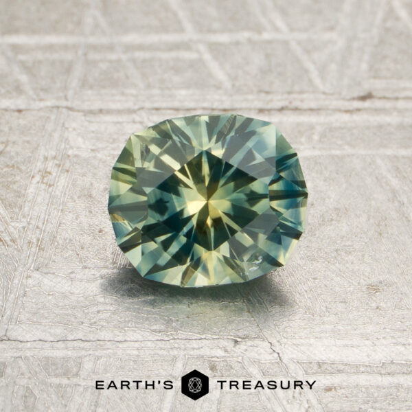 1.01-Carat Teal Green-Yellow Particolored Montana Sapphire (Heated) A 1.01-carat, faceted, round teal green-yellow particolored Montana sapphire (heated) is displayed on a textured, metallic surface. The text “EARTH’S TREASURY” appears at the bottom.