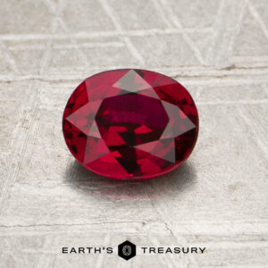 A 1.20-carat heated Mozambique ruby, expertly cut in an oval shape, is displayed on a textured surface. The “EARTH’S TREASURY” logo is visible at the bottom of the image.
