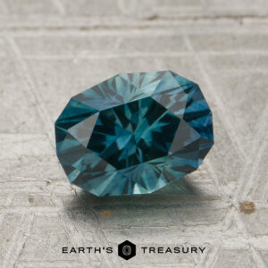 A 1.65-carat teal Montana sapphire (heated) rests on a textured gray surface, with the “EARTH’S TREASURY” logo and text displayed below the stone.