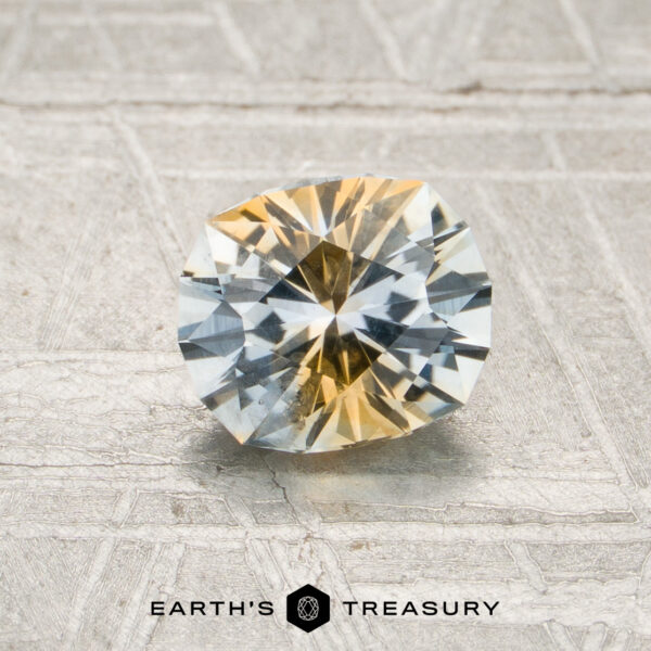 0.98-Carat Colorless-Yellow Particolored Montana Sapphire (Heated) A 0.98-carat colorless-yellow particolored Montana sapphire (heated) rests on a textured surface above the “Earth’s Treasury” logo.