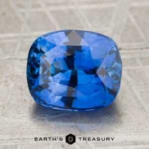A 3.29-carat rich cornflower blue Ceylon sapphire, cut in a cushion shape with faceted surfaces, rests on a textured, light-colored background. The Earth’s Treasury logo appears at the bottom.