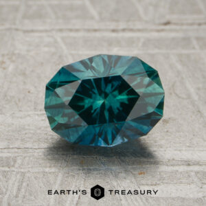 A 1.59-carat Spruce Green Montana Sapphire, expertly oval-cut with intricate facets, is displayed on a textured gray surface, with the text “EARTH’S TREASURY” at the bottom.