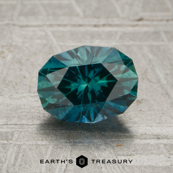 1.59-Carat Spruce Green Montana Sapphire (Heated) A 1.59-carat Spruce Green Montana Sapphire, expertly oval-cut with intricate facets, is displayed on a textured gray surface, with the text “EARTH’S TREASURY” at the bottom.