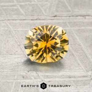 A 0.97-carat golden yellow Montana sapphire (heated) with a cushion cut is displayed on a metallic textured surface, with the “Earth’s Treasury” logo and text at the bottom.