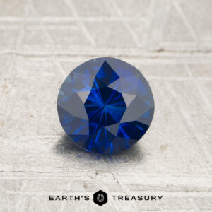 A 1.13-carat royal blue Ceylon sapphire (heated) in a round brilliant cut rests on a textured surface, with “Earth’s Treasury” displayed below.