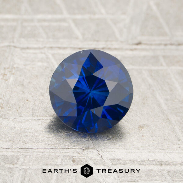 1.13-Carat Royal Blue Ceylon Sapphire (Heated) A 1.13-carat royal blue Ceylon sapphire (heated) in a round brilliant cut rests on a textured surface, with “Earth’s Treasury” displayed below.