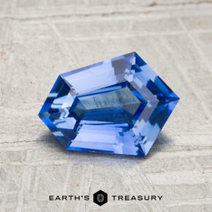 A 1.50-carat rich cornflower blue Ceylon sapphire (heated), cut in a hexagonal shape, rests on a textured gray surface. The “Earth’s Treasury” logo and text are displayed at the bottom of the image.