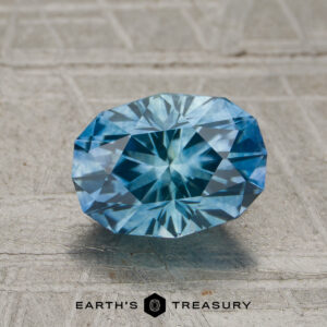 A 1.57-carat Robin’s Egg Blue Montana sapphire is displayed on a textured surface, with the words “Earth’s Treasury” and a hexagonal logo beneath it.