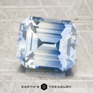 A rectangular, emerald-cut 3.32-carat colorless-sky blue particolored Ceylon sapphire is displayed on a textured gray surface with the “EARTH’S TREASURY” logo below.