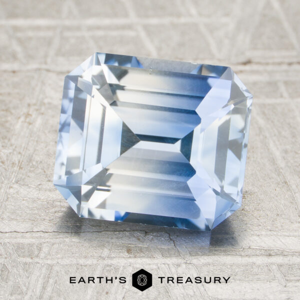 A rectangular, emerald-cut 3.32-carat colorless-sky blue particolored Ceylon sapphire is displayed on a textured gray surface with the “EARTH’S TREASURY” logo below.