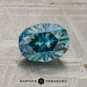 A 1.53-carat heated Montana sapphire with a blue-green faceted oval shape rests on a textured gray surface, with the text “EARTH’S TREASURY” below it.