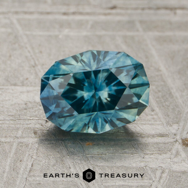 1.53-Carat Teal Montana Sapphire (Heated) A 1.53-carat heated Montana sapphire with a blue-green faceted oval shape rests on a textured gray surface, with the text “EARTH’S TREASURY” below it.
