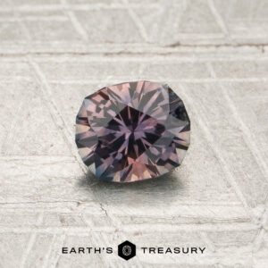 A 0.72-carat mauve-purple particolored Montana sapphire (heated), cut in a faceted round shape with hues of purple and dusty pink, is displayed on a textured, light-colored surface. The “Earth’s Treasury” logo appears at the bottom.