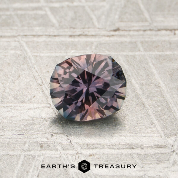 0.72-Carat Mauve-Purple Particolored Montana Sapphire (Heated) A 0.72-carat mauve-purple particolored Montana sapphire (heated), cut in a faceted round shape with hues of purple and dusty pink, is displayed on a textured, light-colored surface. The “Earth’s Treasury” logo appears at the bottom.