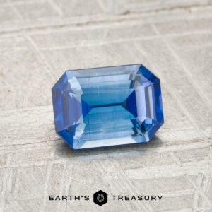 A 1.27-carat medium blue Ceylon sapphire (heated), cut in a rectangular faceted shape, rests on a textured light surface, with the words “EARTH’S TREASURY” and a logo at the bottom.