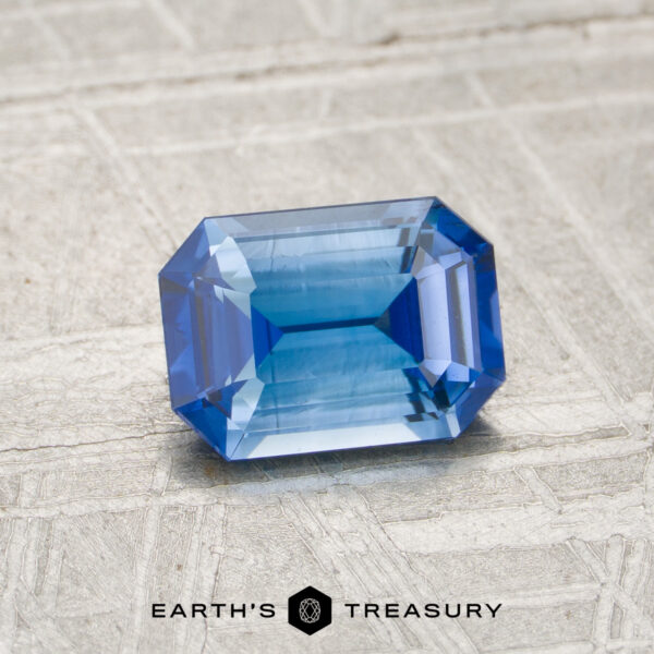 1.27-Carat Medium Blue Ceylon Sapphire (Heated) A 1.27-carat medium blue Ceylon sapphire (heated), cut in a rectangular faceted shape, rests on a textured light surface, with the words “EARTH’S TREASURY” and a logo at the bottom.
