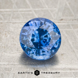 A 2.50-carat rich cornflower blue Ceylon sapphire (heated) in a round brilliant cut, displayed on a textured surface with the “EARTH’S TREASURY” logo and text underneath.