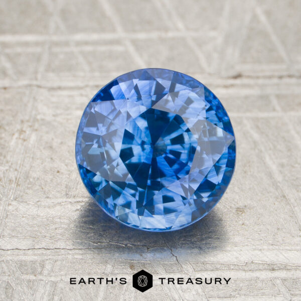A 2.50-carat rich cornflower blue Ceylon sapphire (heated) in a round brilliant cut, displayed on a textured surface with the “EARTH’S TREASURY” logo and text underneath.