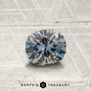 A 0.72-carat steely blue-gray particolored Montana sapphire (heated), cut in an oval shape with a faceted surface, is displayed on a textured metallic background, featuring the “Earth’s Treasury” logo at the bottom.
