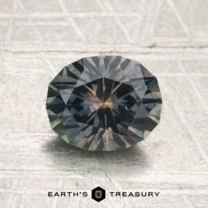 A round, faceted 1.51-carat gray Montana sapphire with dark blue and green hues sits on a textured surface, with “EARTH’S TREASURY” written below the gem.