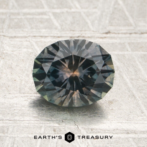 1.51-Carat Gray Montana Sapphire (Heated) A round, faceted 1.51-carat gray Montana sapphire with dark blue and green hues sits on a textured surface, with “EARTH’S TREASURY” written below the gem.
