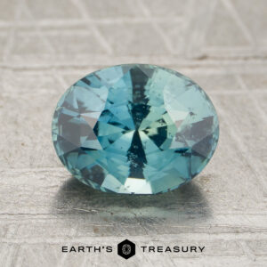 A 1.89-carat teal Madagascar sapphire, cut in an oval shape, is displayed on a textured surface with the text “EARTH’S TREASURY” and a hexagon logo at the bottom.