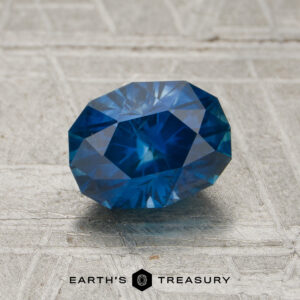 A 1.32-carat rich blue Montana sapphire (heated) is displayed on a textured gray surface with the “Earth’s Treasury” logo below it.