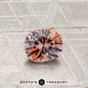 A 0.70-carat orange-lilac particolored Montana sapphire (heated) with a round cut is displayed on a textured silver surface, with the text “EARTH’S TREASURY” at the bottom of the image.