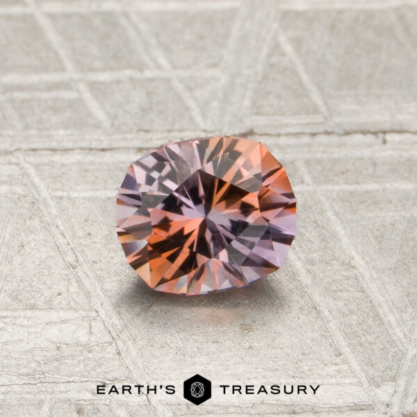 A 0.70-carat orange-lilac particolored Montana sapphire (heated) with a round cut is displayed on a textured silver surface, with the text “EARTH’S TREASURY” at the bottom of the image.
