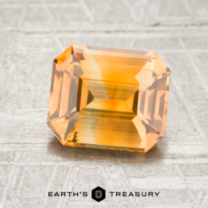 A 2.26-carat orange-yellow particolored sapphire, expertly faceted in a rectangular shape, rests on a textured surface featuring the “Earth’s Treasury” text and hexagonal logo below it.