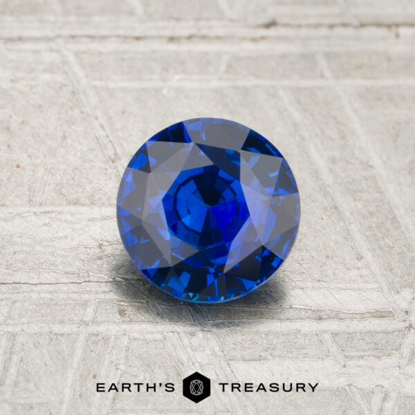 1.05-Carat Royal Blue Ceylon Sapphire (Heated) A 1.05-carat royal blue Ceylon sapphire (heated), round brilliant-cut, is showcased on a light textured surface with the “EARTH’S TREASURY” logo and text at the bottom.