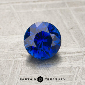 A 1.12-carat royal blue Ceylon sapphire (heated), cut in a round brilliant shape, is displayed on a textured surface with the text “EARTH’S TREASURY” below.