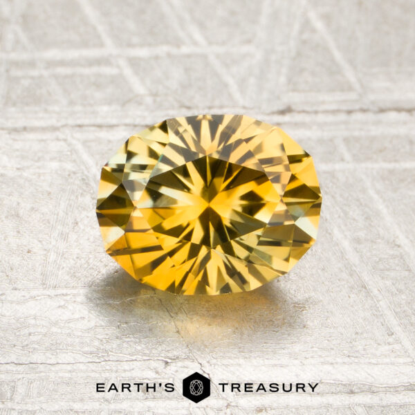 1.45-Carat Golden Yellow Montana Sapphire (Heated) A 1.45-carat golden yellow Montana sapphire (heated), oval-cut, showcased on a textured light surface with the “EARTH’S TREASURY” name and hexagon logo beneath.