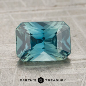 A 2.04-carat Teal Madagascar Sapphire with a rectangular faceted cut is displayed on a textured, light-colored surface. The “EARTH’S TREASURY” logo appears at the bottom.