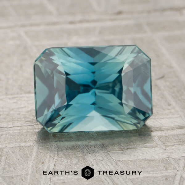 2.04-Carat Teal Madagascar Sapphire (Unheated) A 2.04-carat Teal Madagascar Sapphire with a rectangular faceted cut is displayed on a textured, light-colored surface. The “EARTH’S TREASURY” logo appears at the bottom.