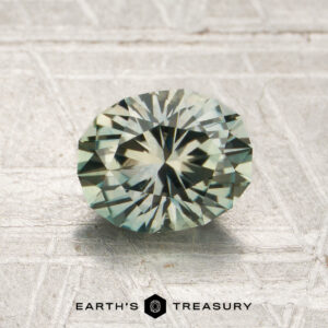 1.39-carat pale grayish green Montana sapphire (heated), oval-cut and faceted, displayed on a light textured surface, with the text “Earth’s Treasury” below.