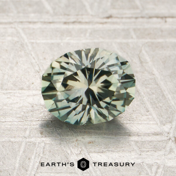 1.39-Carat Pale Grayish Green Montana Sapphire (Heated) 1.39-carat pale grayish green Montana sapphire (heated), oval-cut and faceted, displayed on a light textured surface, with the text “Earth’s Treasury” below.